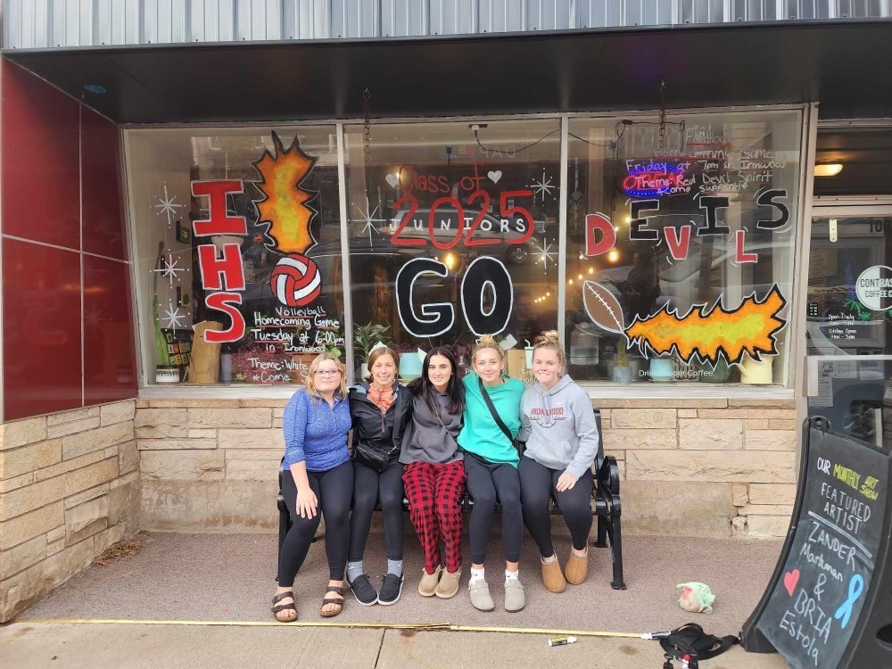 5 students sit in front of a store window with homecoming decorations